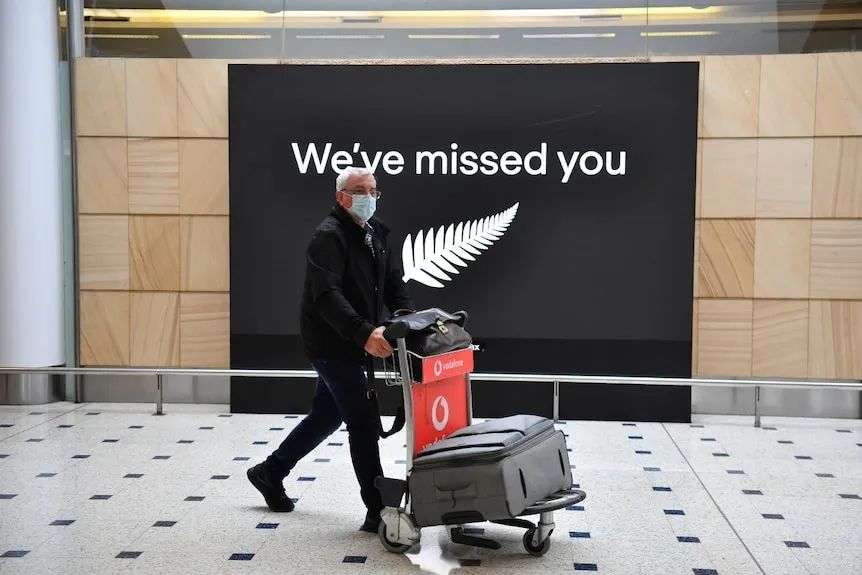 A man in a mask pushes a suitcase trolley at Sydney airport, in front of a sign with silver fern that says: 