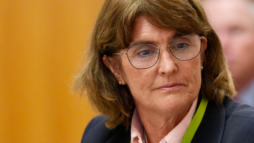 A woman with short brown hair and glasses looks down slightly.