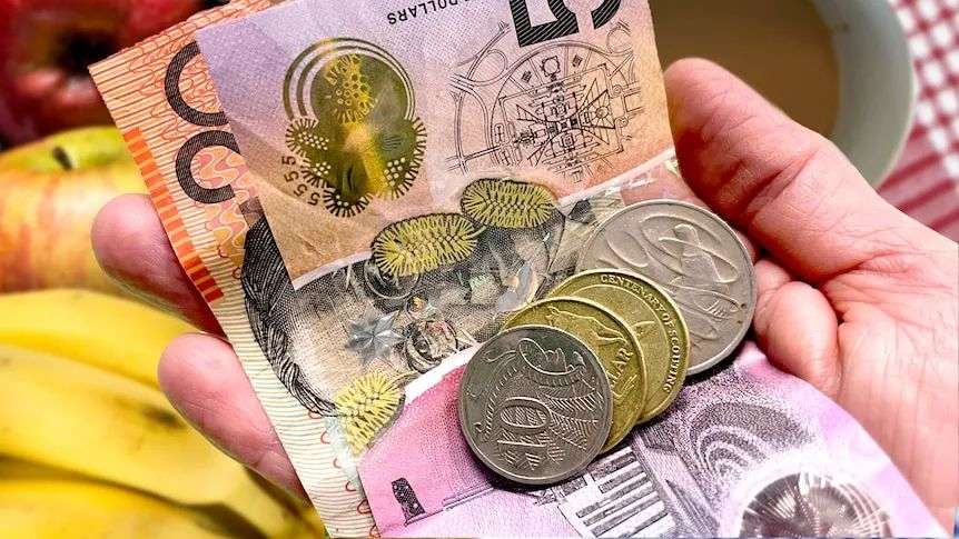 Hand holding money, notes and coins with a coffee cup and fruit in the background, cost of living