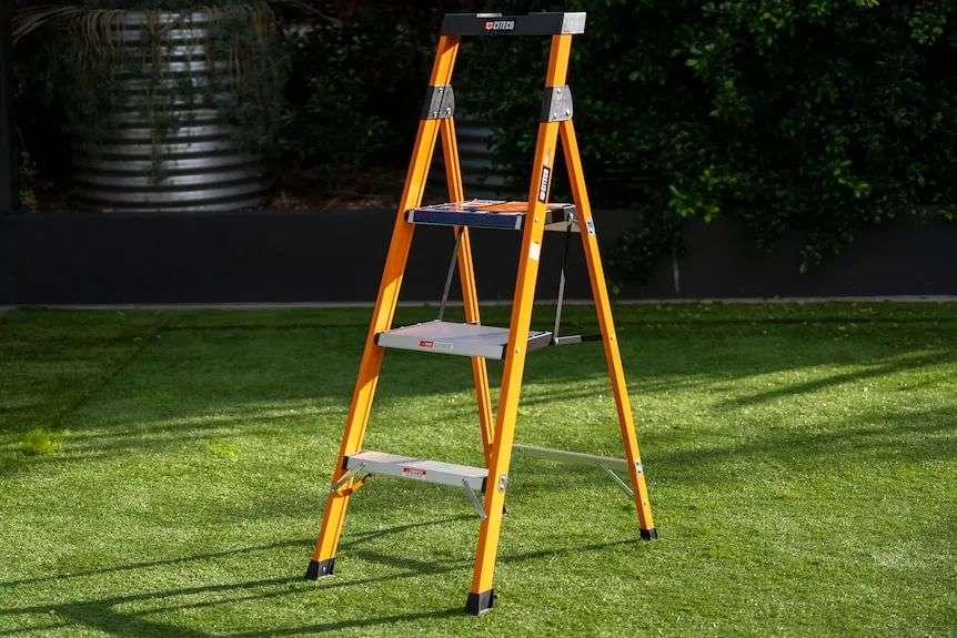 An orange 3-step ladder stands on a patch of fake turf, in the sun outdoors.