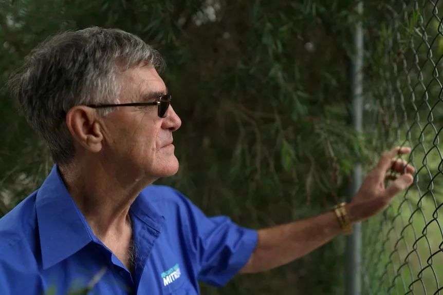 A man looks ahead as he reaches out and holds a wire fence. His expression is neutral.