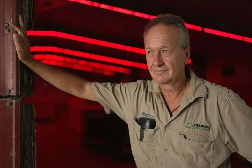 A man leans against a wall in a darkened rim lit only by strips of red light. He is wearing a work shirt has a slight smile.