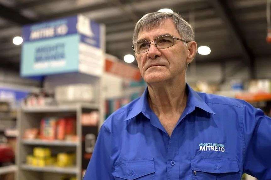 A man in a mitre 10 shirt looks ahead with a sad expression. He is standing a hardware store.