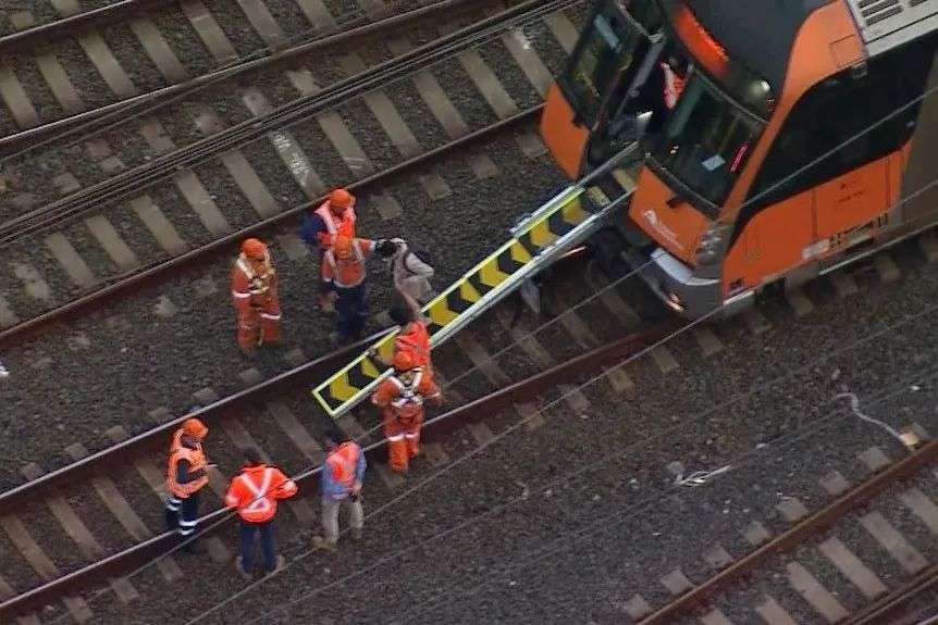 Crews help people off orange train carriage down yellow-and-black plank after overhead wire fell on carriages