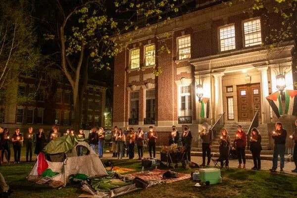 Students standing in a circle around a tent draped with a Palestinian flag hold candles near a campus building.