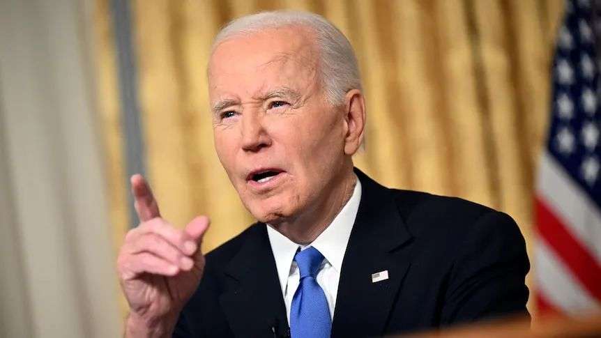 An elderly man in a suit holds up his finger as he speaks behind a wooden desk in front of an American flag.