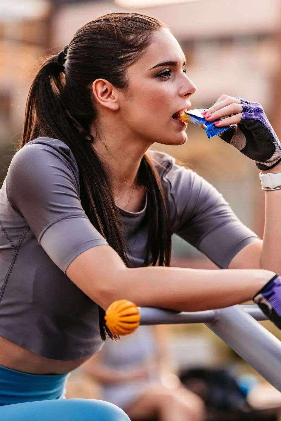Young beautiful athlete woman training on outdoors exercise bike in park and eating energy bar.
