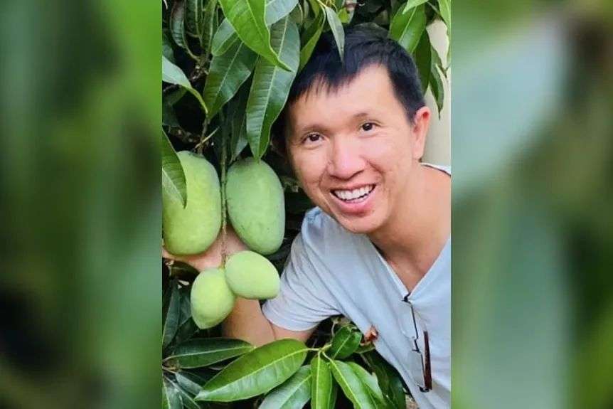 A man poses smiling next to some mangoes on a tree.