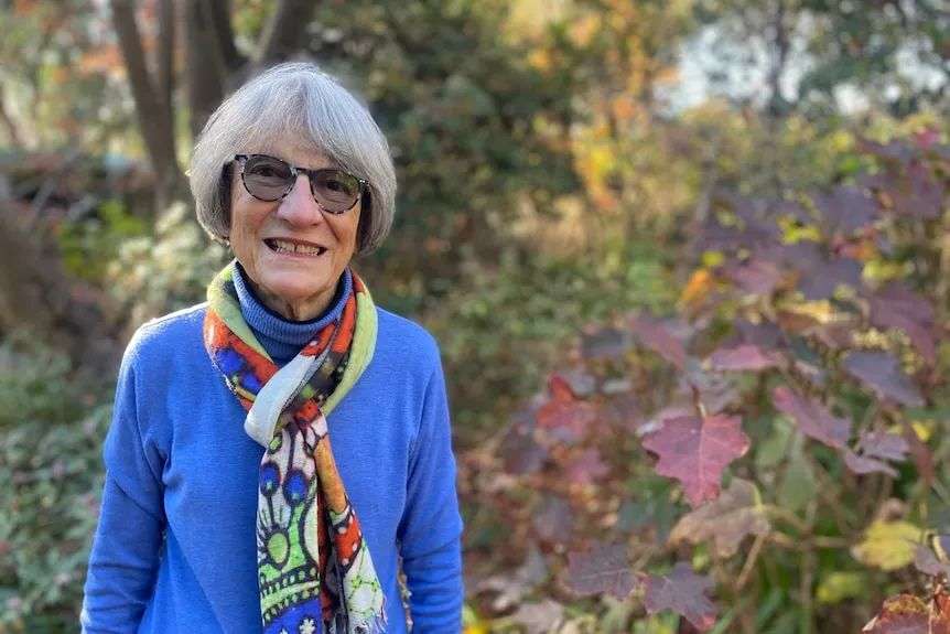 Bespectacled woman with grey bob of hair, blue pullover and scarf standing in her garden