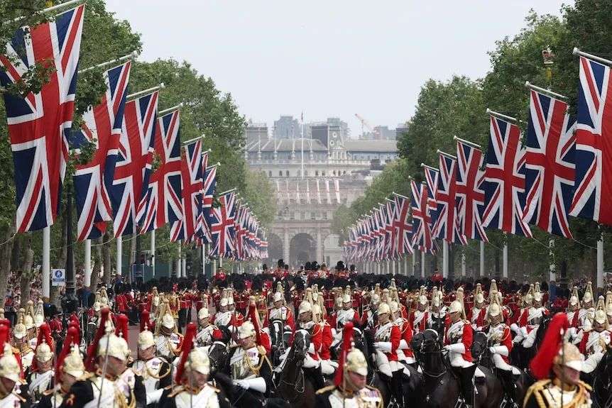 A wide shot of troops in ceremonial dress taking part in a parade. The street is lined with Union Jack flags.