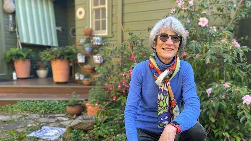 Bespectacled woman with grey bob of hair, blue pullover and scarf in front of her cottage