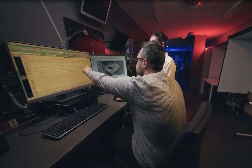 Two please examine two computer screens, one showing brainwaves, the other a camera view of the sleeper.