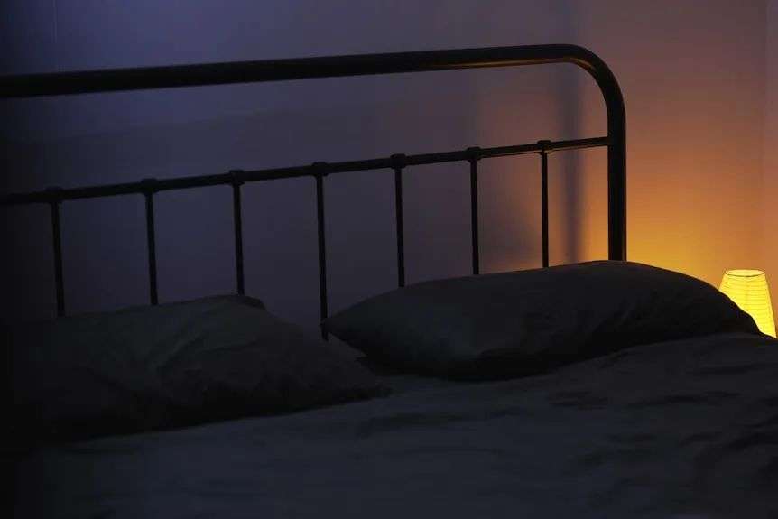 A black bed frame with dark sheets and a small table lamp nearby lit up in darkness.