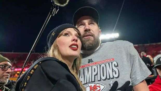 Taylor Swift wearing a black hat and hoodie posing with Travis Kelce after a championship game