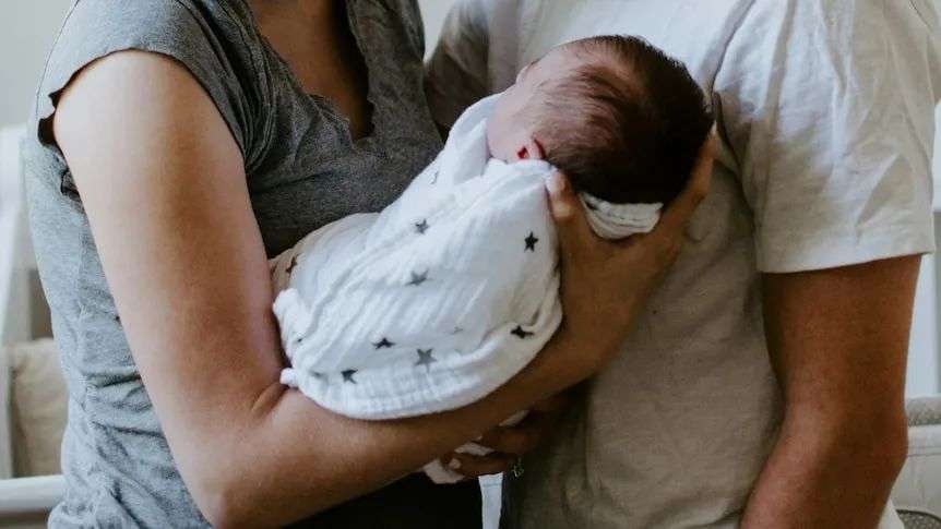 A close up of a baby in white muslin wrap with stars on it, being held by two adults. No faces are visible.