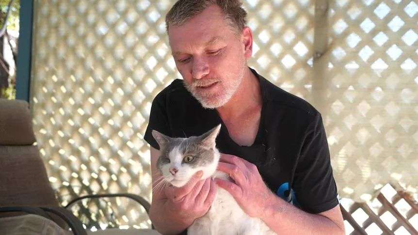 A man in a black T-shirt affectionately holds his one-eyed cat under the chin