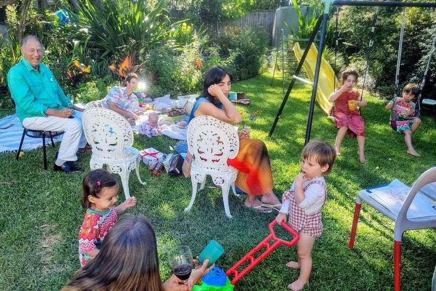 family sit in the backyard on chairs and blankets with drinks and kids on swings