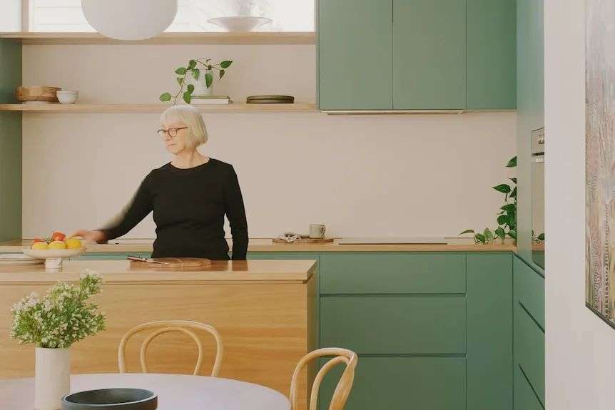 A woman stands behind a timber bench in a bright modern kitchen.