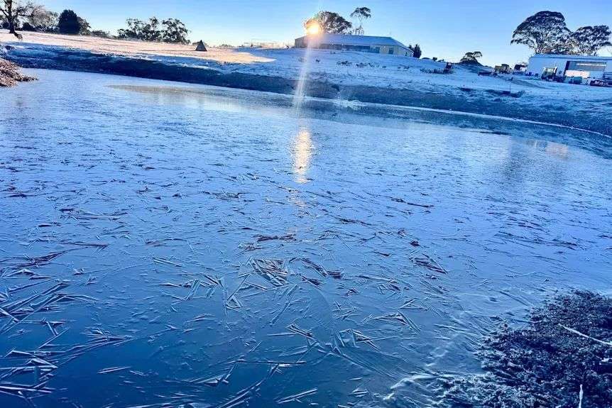 The sun rises over a frozen lake on frost-covered banks with a house covered in white in the background
