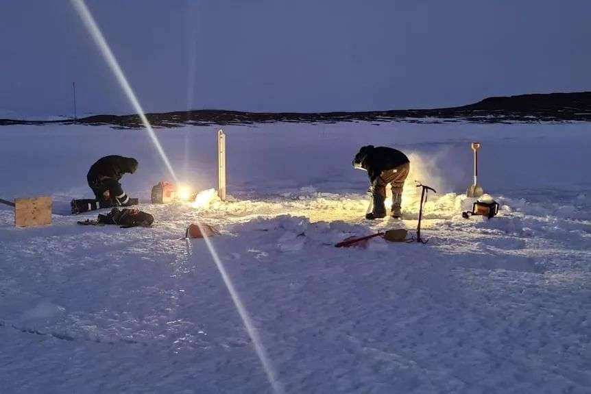 Two people cut a hole in the ice by torchlight at Mawson station, Antarctica, June 2023