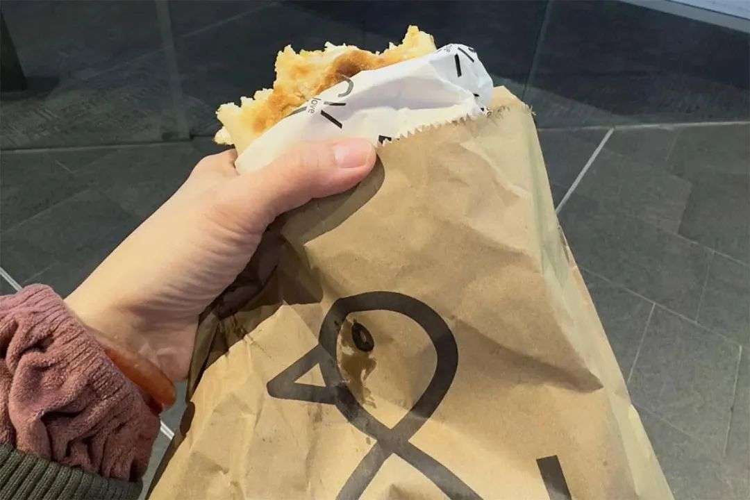 A woman holds a brown paper bag with a bacon and egg roll inside that is half eaten.