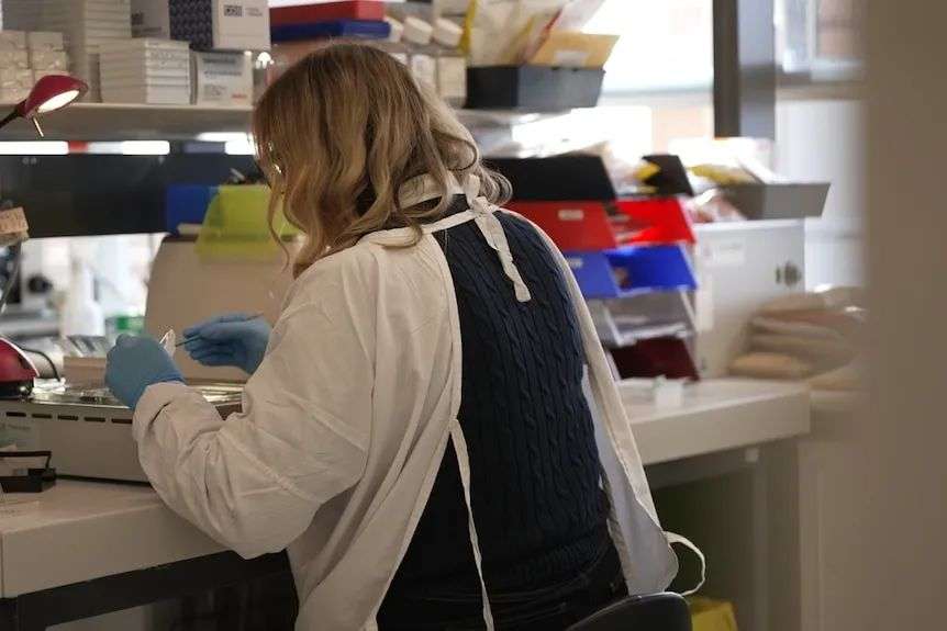 A woman wearing a white lab coat over a navy jumper sits in a laboratory handling samples.