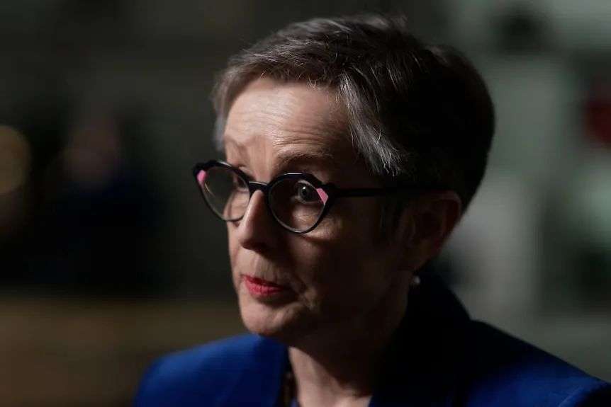 A woman with short grey hair, rounded black glasses and a blue blazer sitting in an office.