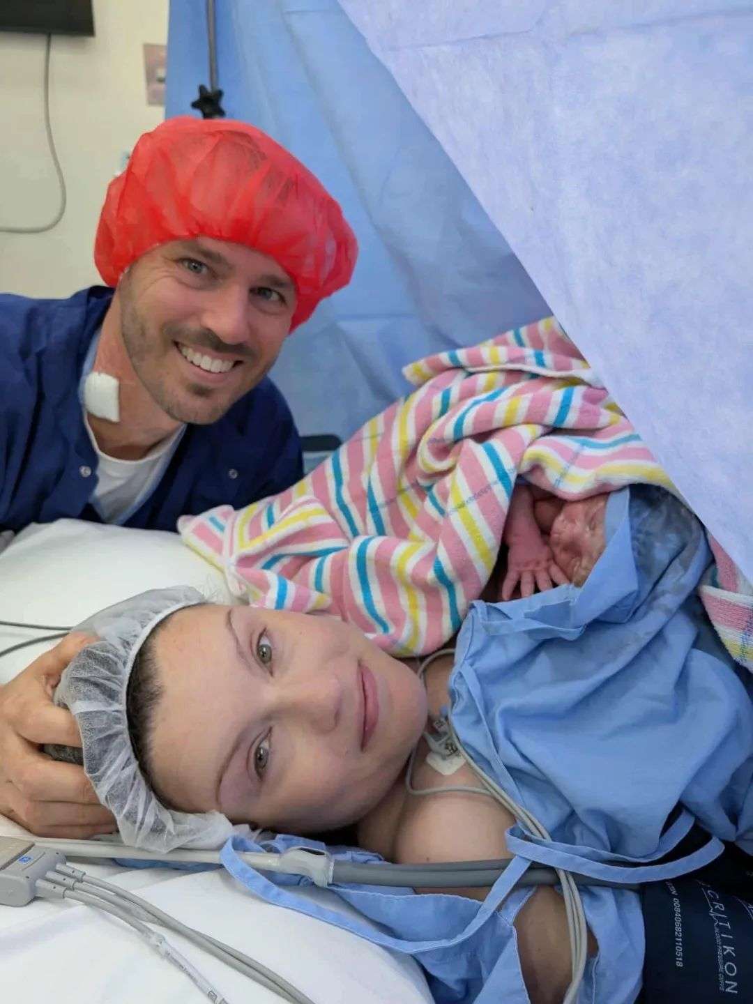 A woman lying on a hospital bed holds a newborn baby. A man wearing a hairnet smiles behind them.