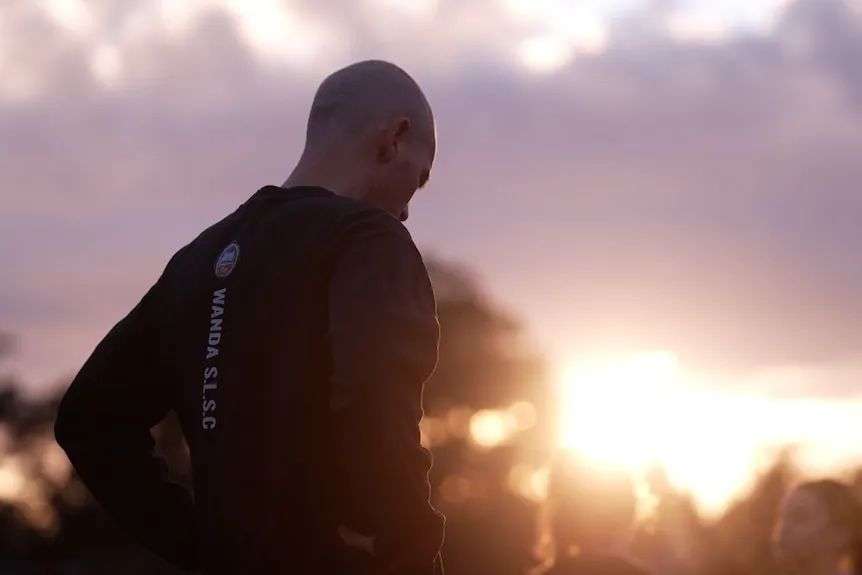 A man is seen from behind wearing a long-sleeved black tshirt and standing with his hands on his hips as the sun rises.