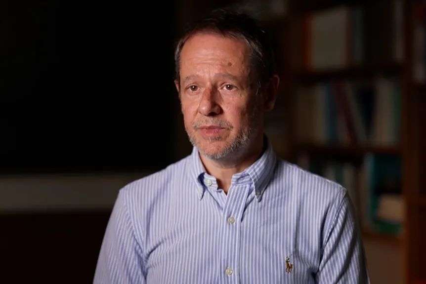 A man in a blue and white vertical striped business shirt sitting in a dark office in front of a bookshelf.