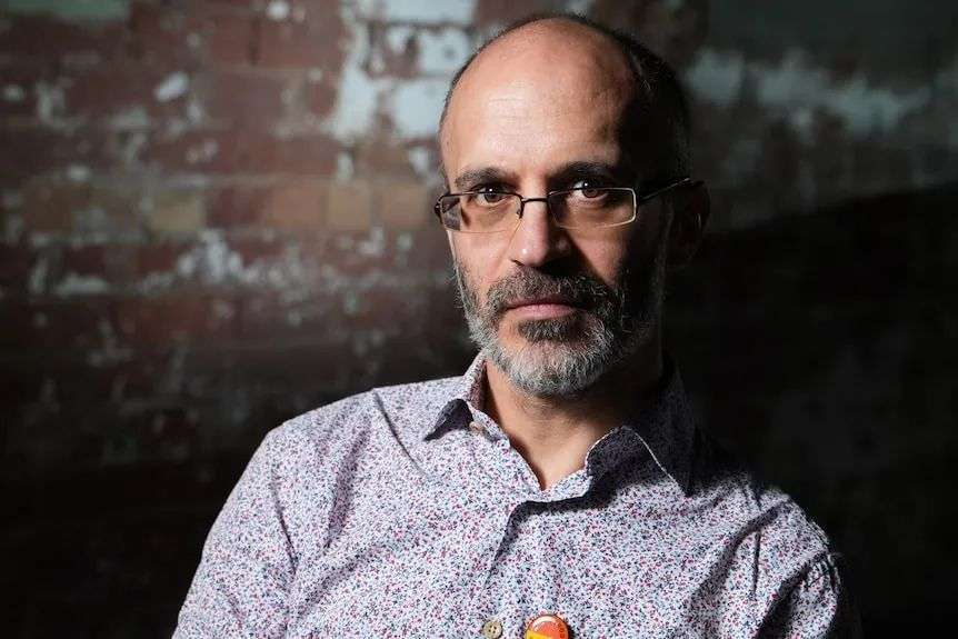 A man with frameless glasses, greying beard and paisley business shirt in front of a brick wall.