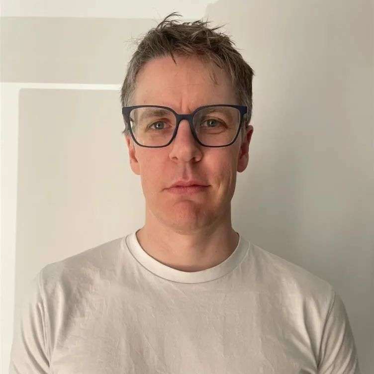 A young man with blonde hair, a grey tshirt  and black glasses stares at the camera, standing in front of a white background.