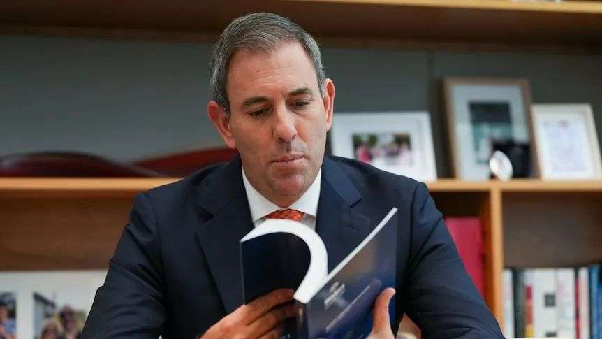 Jim Chalmers reads the budget book in his office, wears blue suit, red tie, shelf behind him as photo frames.