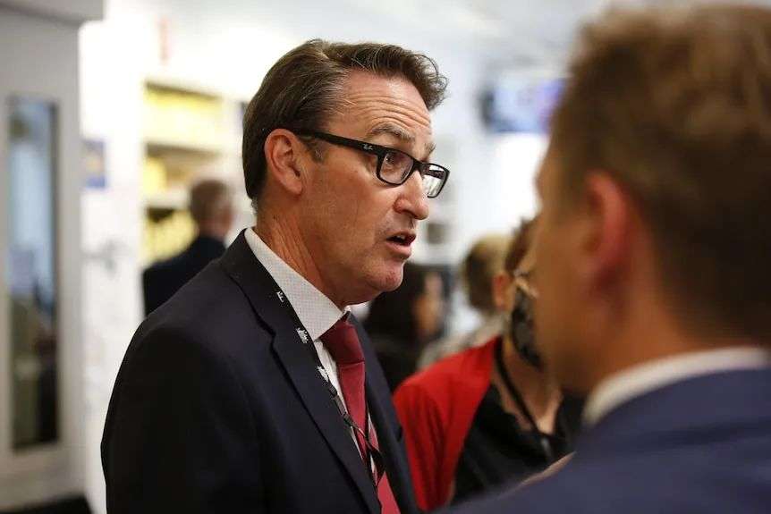 Man in black suit, red tie, black-rimmed glasses, lanyard around neck talks to other not fully in the picture.