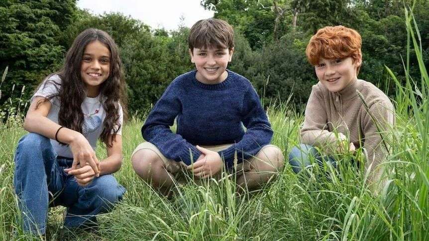 Three children crouch in long grass and smile