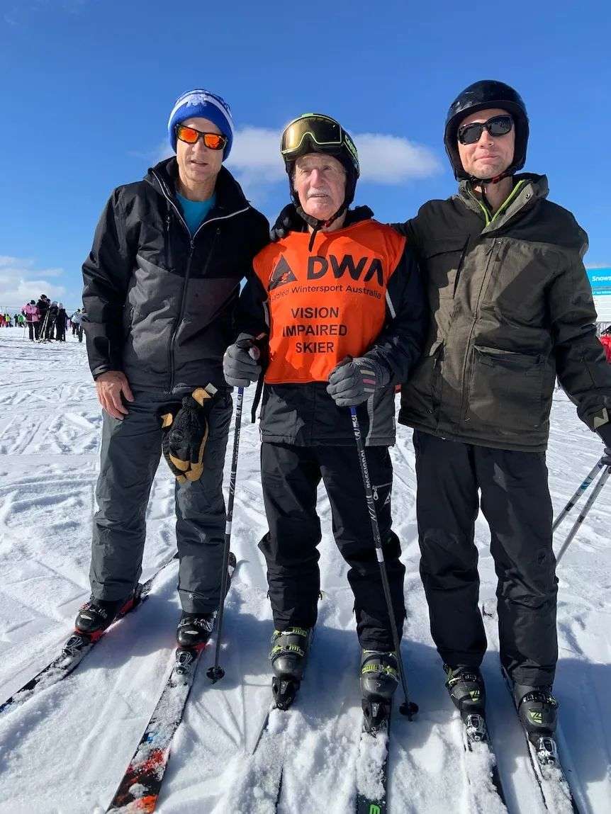 Three men stand in the snow, the elderly man in the middle wears a vest that says he is vision impaired