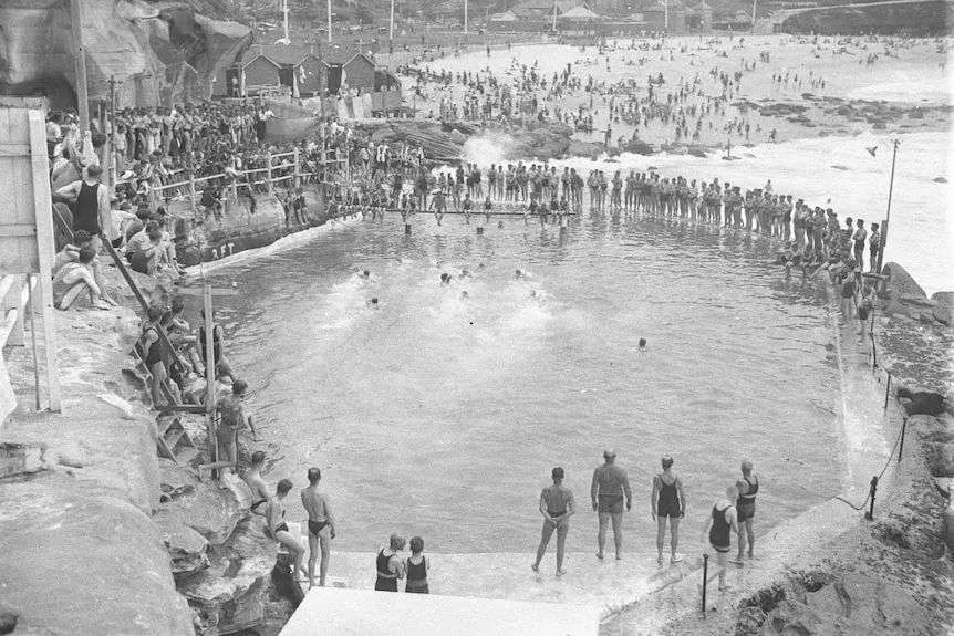 A black and white photo of an ocean pool with people swimming and many others watching.