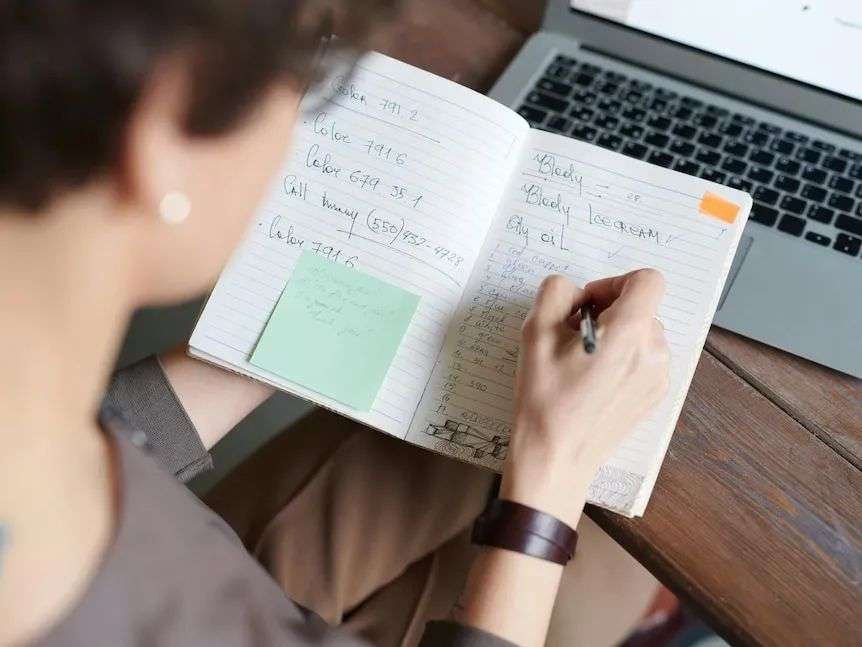 Top view of a person writing a list on her notebook placed against a laptop