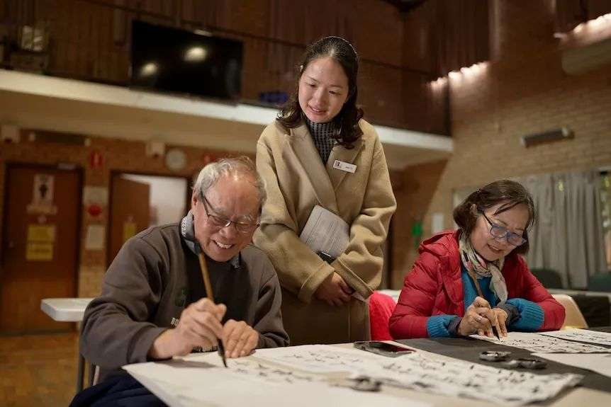 A Chinese woman stands and watches over an elderly man and woman practicing Chinese calligraphy.