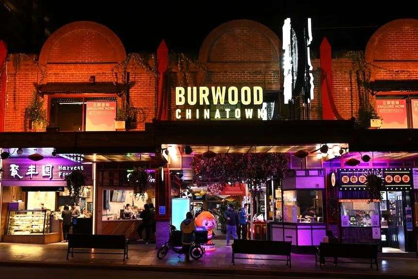 A bustling street with lights at night, with a big Burwood Chinatown sign in the middle of the brick building.