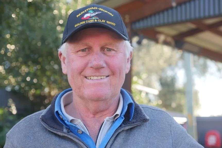 Smiling Caucasian-looking older man, grey hair, wears dark cap with motto, grey, blue jumper over light shirt, blurred behind.