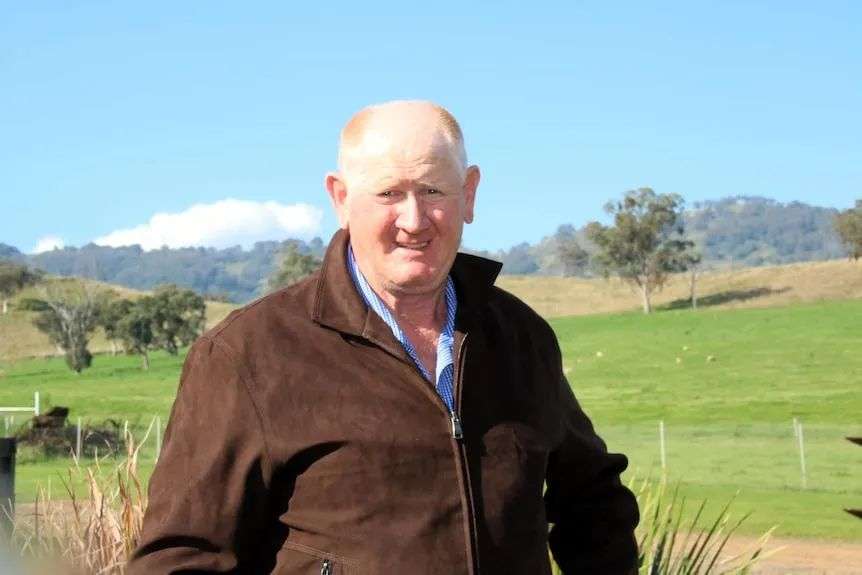 An elderly man with blue cap, blue shirt, brown jacket, looks at camera, neutral expression, porce roof, trees behind.