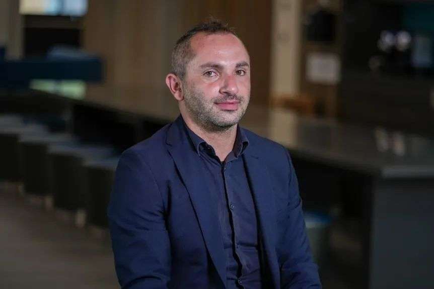 A man with short hair and a short beard sitting in a room. He is wearing a navy shirt and jacket