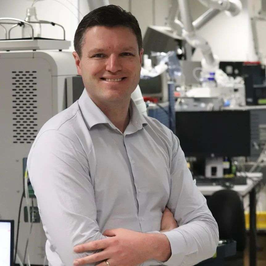 A young man in a business shirt smiles with his arms folded in a lab.