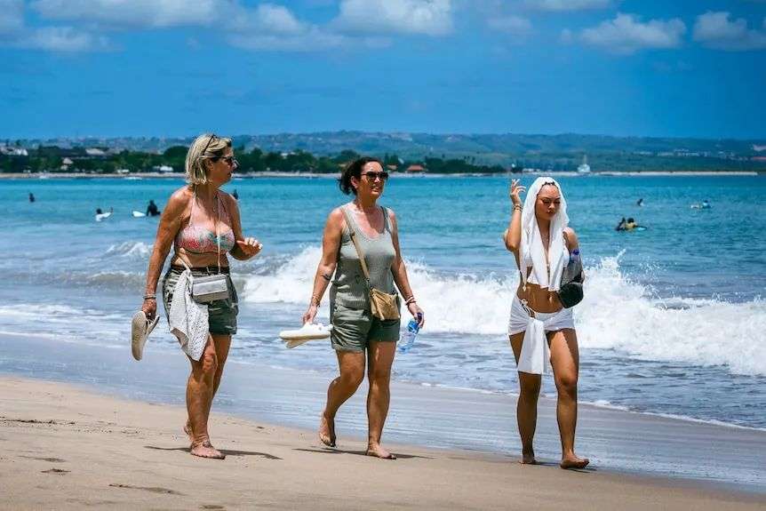 Three girls walk along a white sandy beach on a sunny day.
