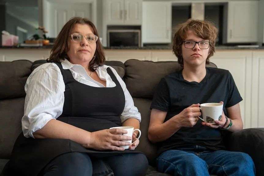 A woman and a teenage boy each holding a mug while sitting on a couch with a kitchen in the background