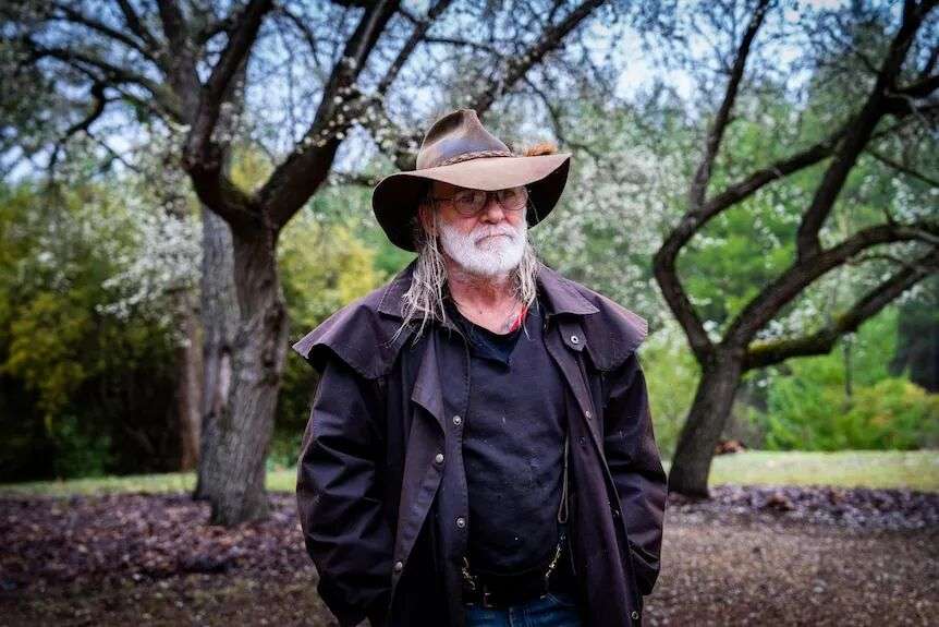 一位身穿黑色衣服、头戴棕色帽子的老人站在树前,双手插在口袋里