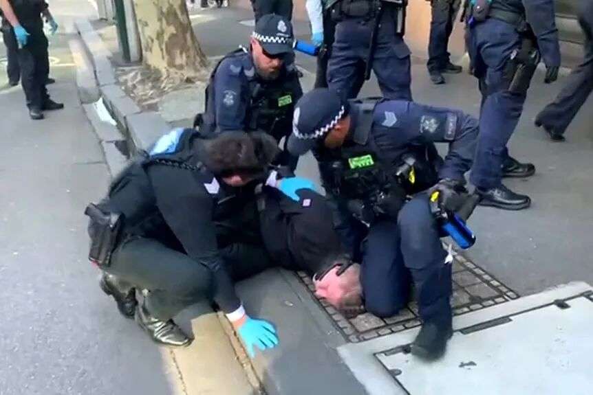 A man with blonde hair is held down on a footpath by several police officers.