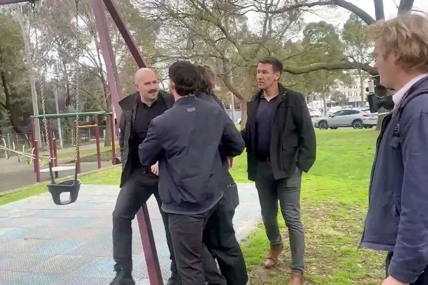 A bald man with a brown moustache is confronted by several men in a playground.