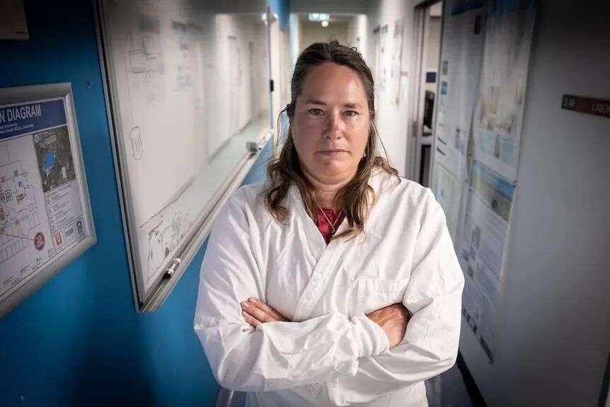Kirsten stands waering a lab coat in the wall walled hallway of a university lab.